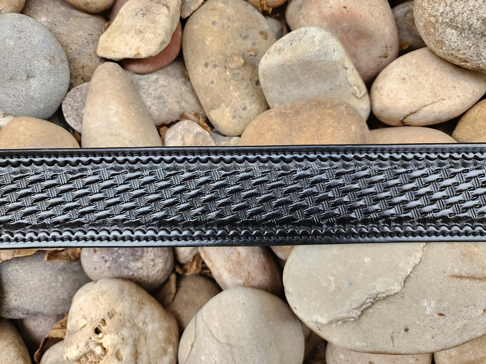 Close up view on the black leather basket weave pattern tooled on the belt, the belt lays on a bed of pale river rocks.
