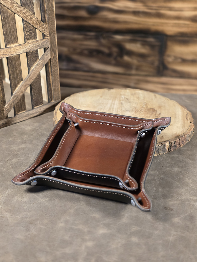 Two brown leather catch-all trays, nested inside each other, on a wooden surface with a rustic background
