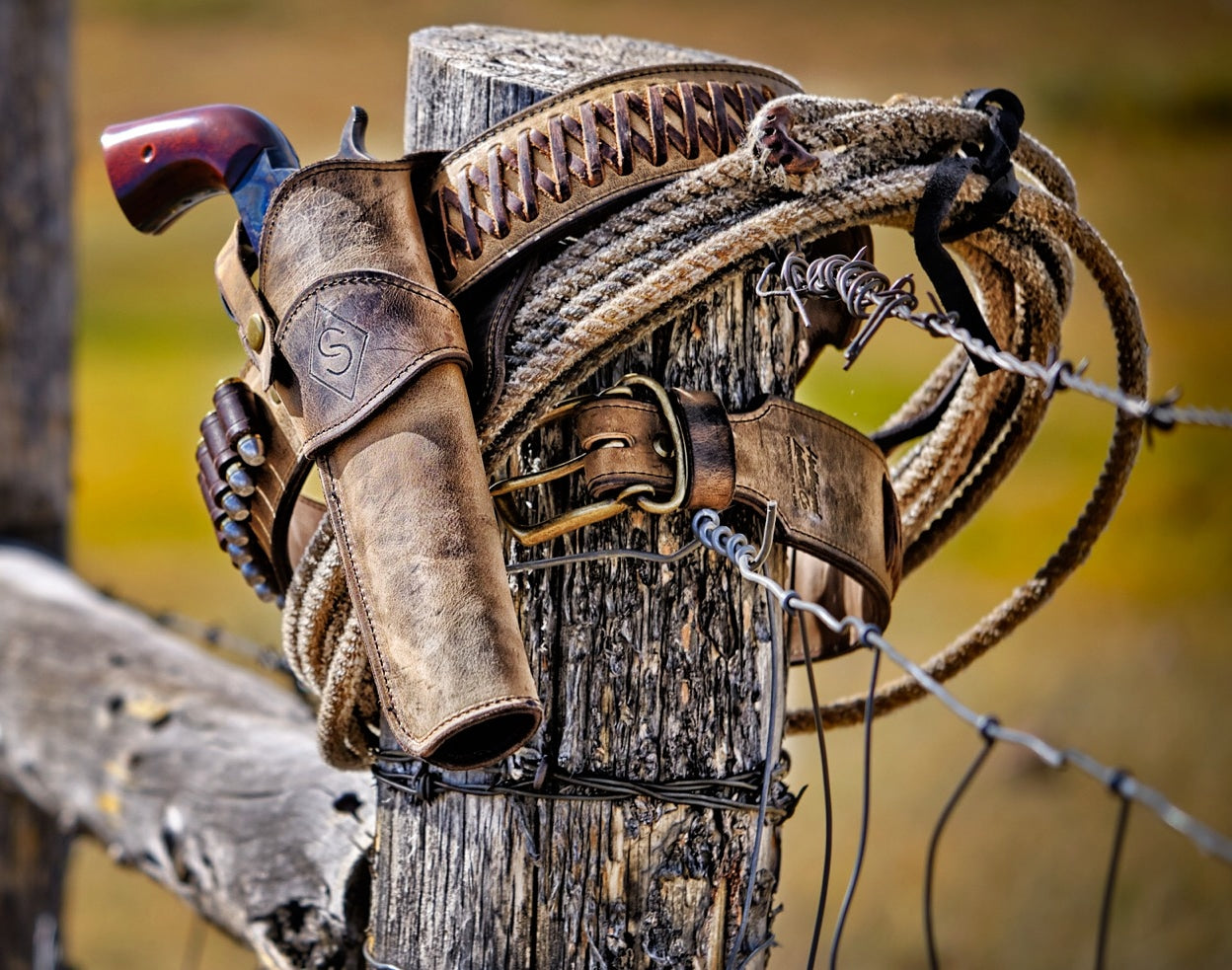 Rustic distressed leather holster and cowboy belt resting on a wooden ranch fencepost with coiled cattle rope and barbed wire.