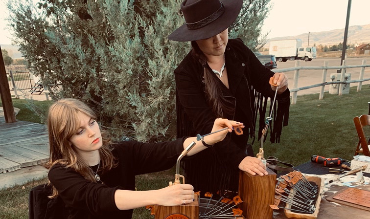 Two women wearing black stand at a table heating mini branding irons to personalize leather gifts for guests at an outdoors event