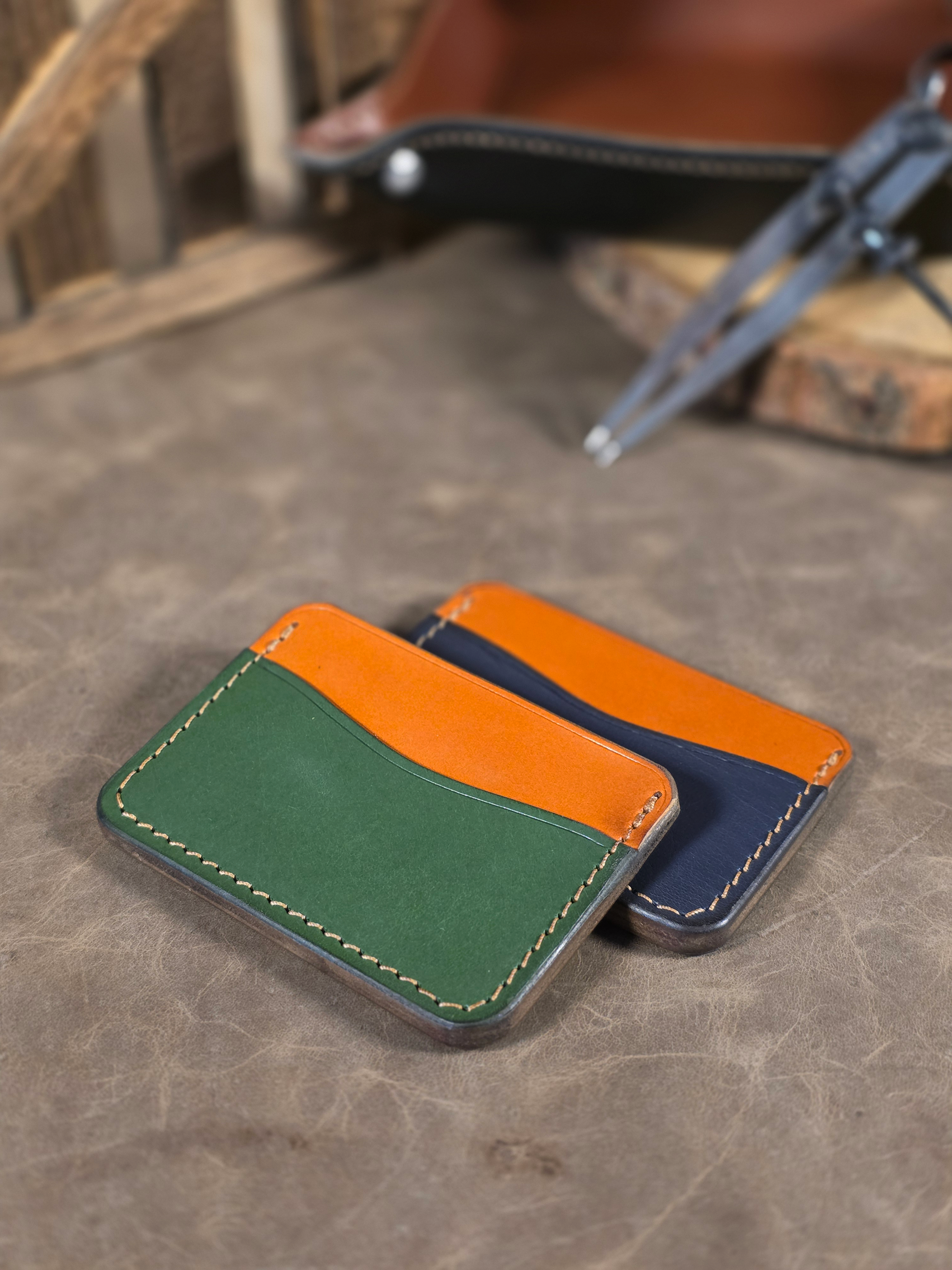 Two minimalist leather card wallets sit staggered on a leather surface, one is tan and navy, the other is tan and green