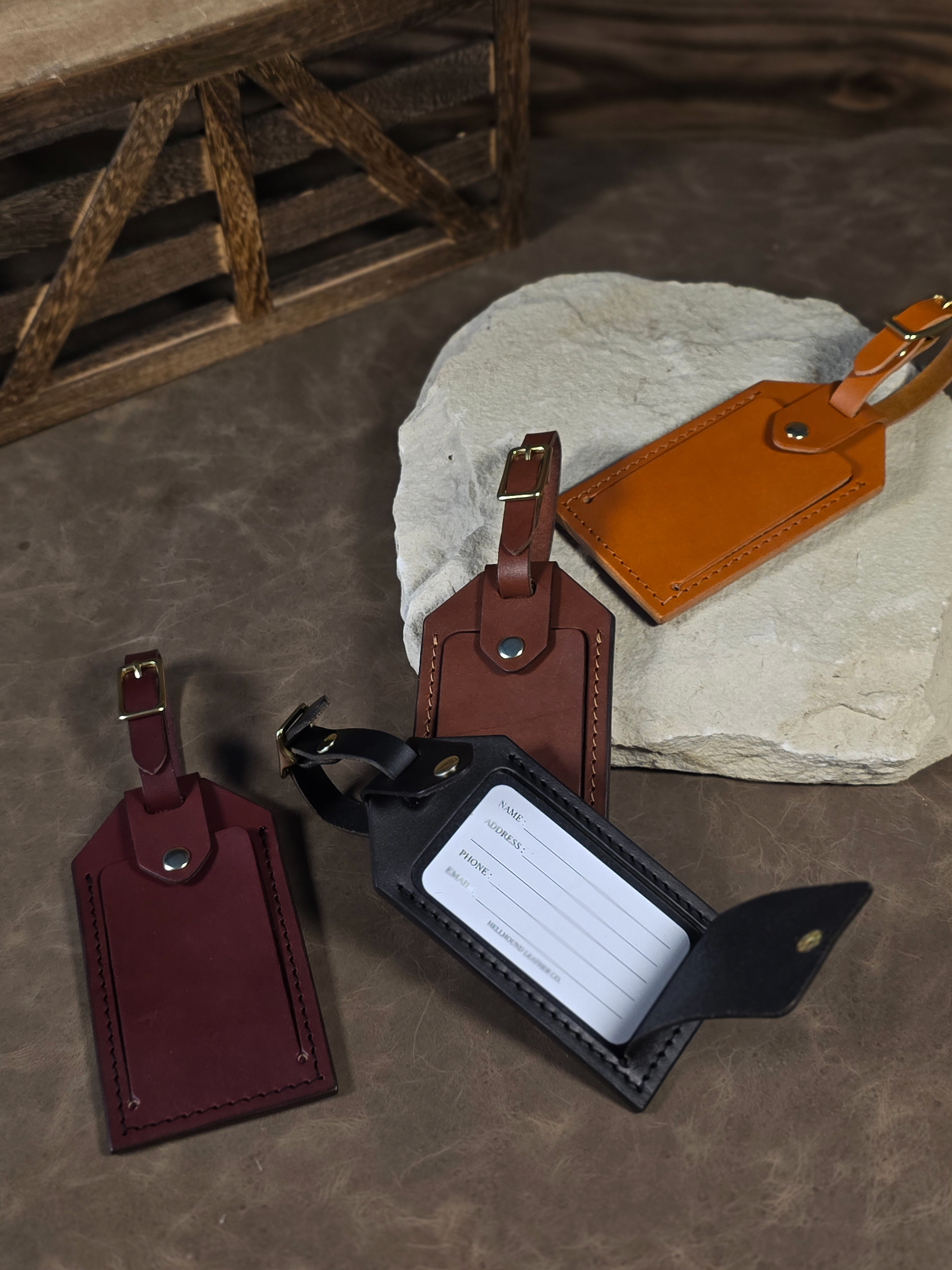 Four leather luggage tags with solid brass snap for opening or closing the window. The four luggage tags are made with russet, oxblood, black, or brown leather. The russet luggage tag sits on a white stone while the others are positioned on the rustic leather background.