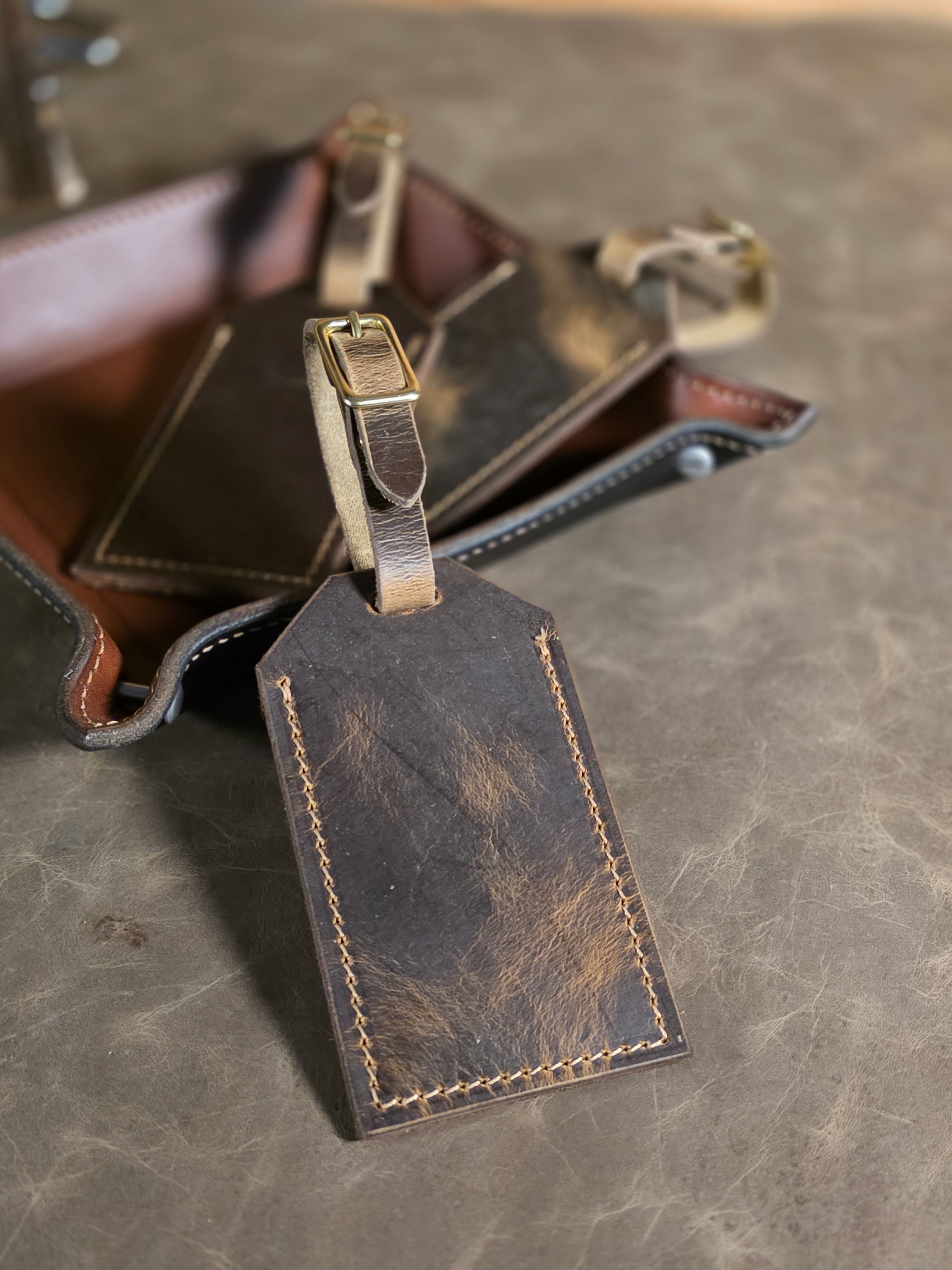 Waxed crazyhorse rustic leather luggage tag with solid brass hardware resting against the edge of a leather valet tray atop a leather mat.