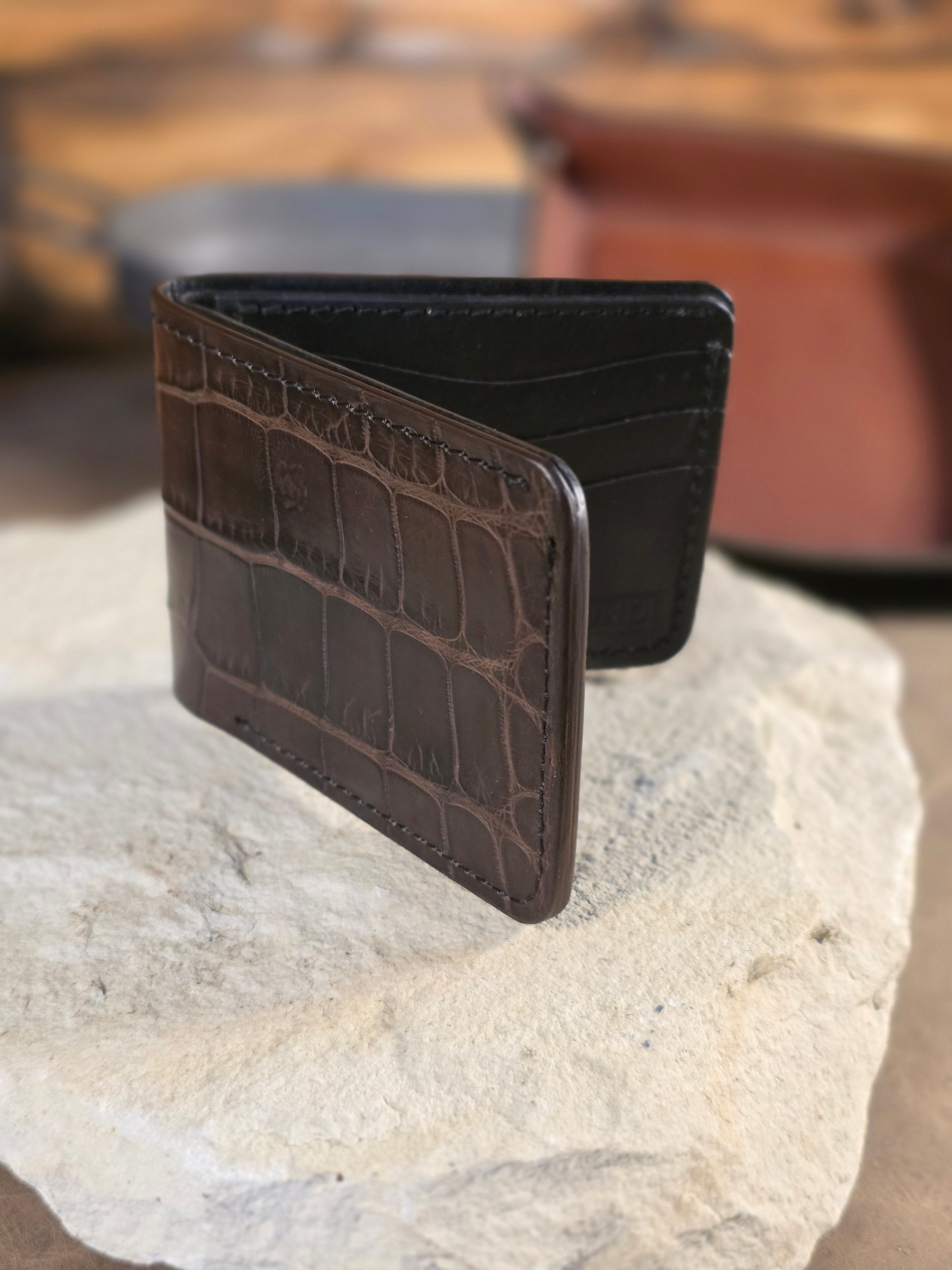 Beautiful deep brown alligator wallet sits propped up, but open, on a white stone base. The black leather interior can be seen in the rear-ground.