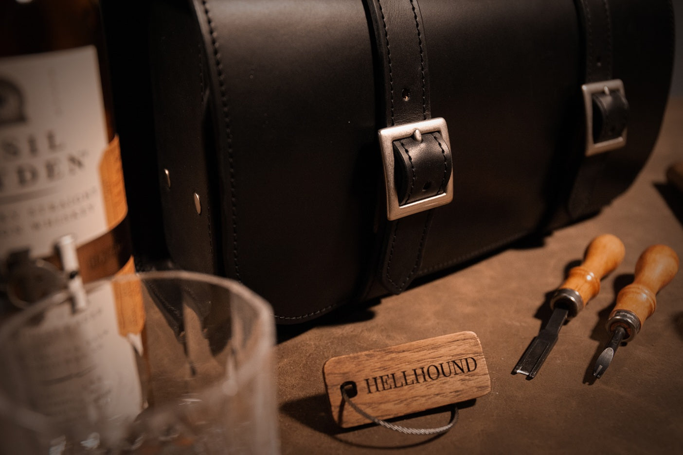 Close-up of a leather bag with tools and a bottle labeled 'Hellhound' on a wooden surface.