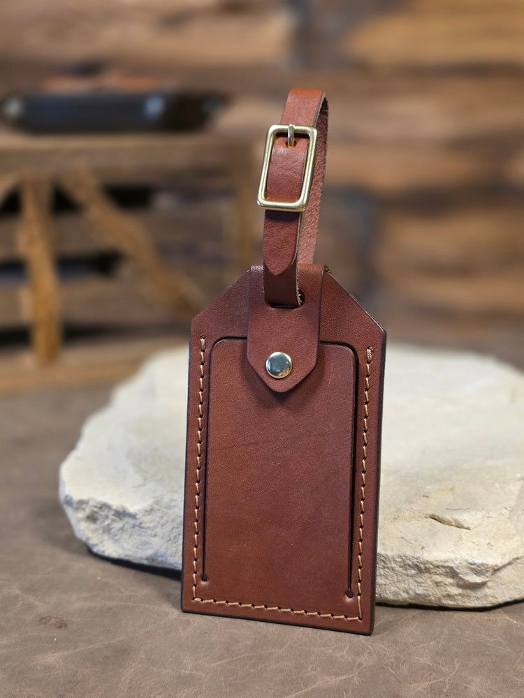 Close up view of a brown leather windowed luggage tag finished with solid brass hardware and tan stitching. The luggage tag leans against a white rock.