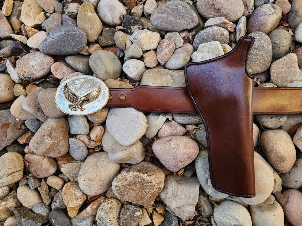 Lone Wolf Mcquade holster, belt, and buckle laying on a bed of pale river rocks. The holster is made from brown leather with black stitching, the belt is made of brown leather as well and the buckle is silver and gold plated.