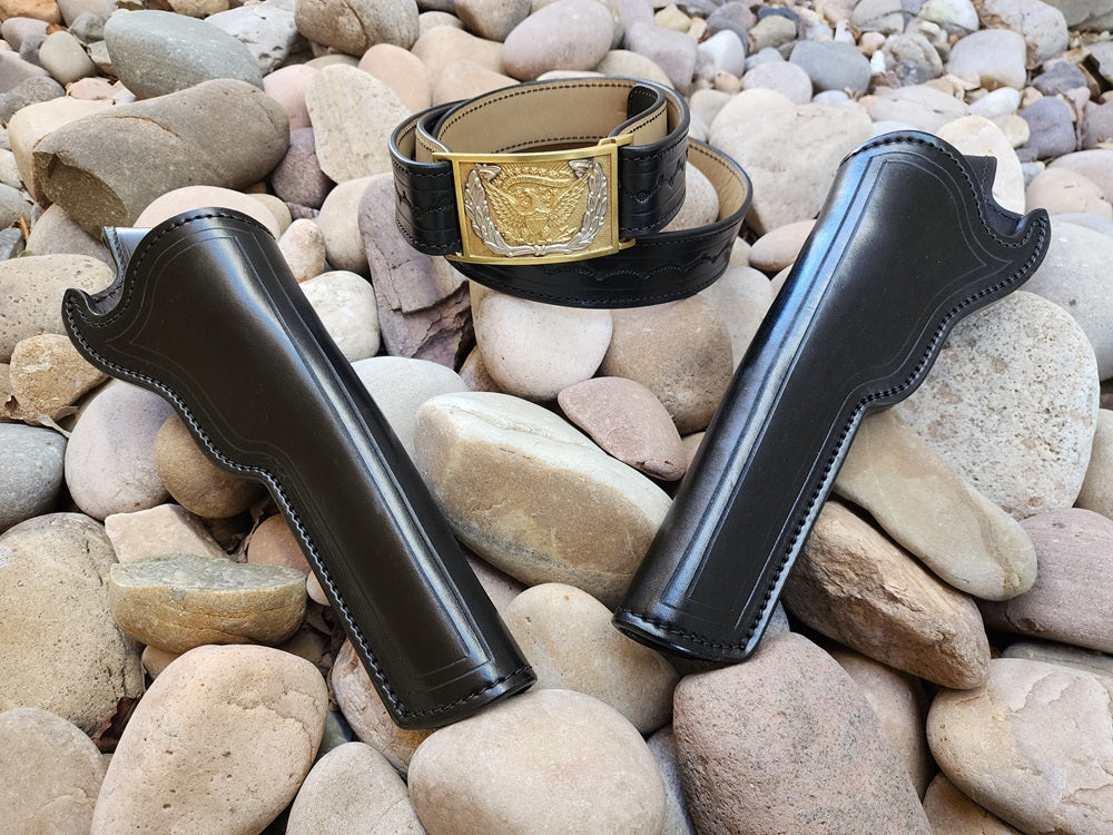 A Black leather replica of Wild Bill Hickok's gun rig. The belt features a traditional silver and gold cavalry officer buckle, the rig itself sits on a bed of pale rocks.