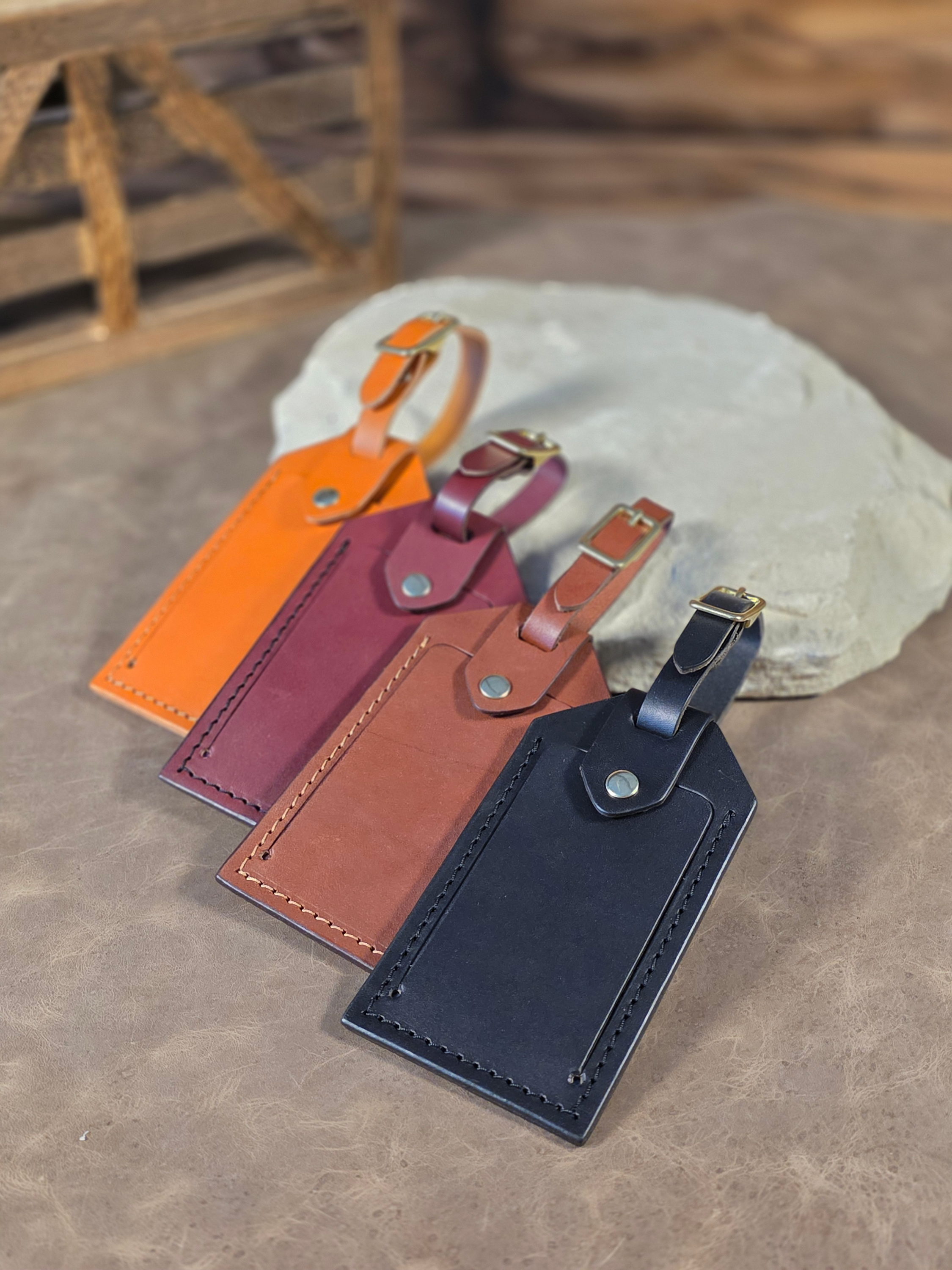 Four leather windowed luggage tags in russet, brown, black, and oxblood coloring finished with solid brass hardware leaning against a white rock.
