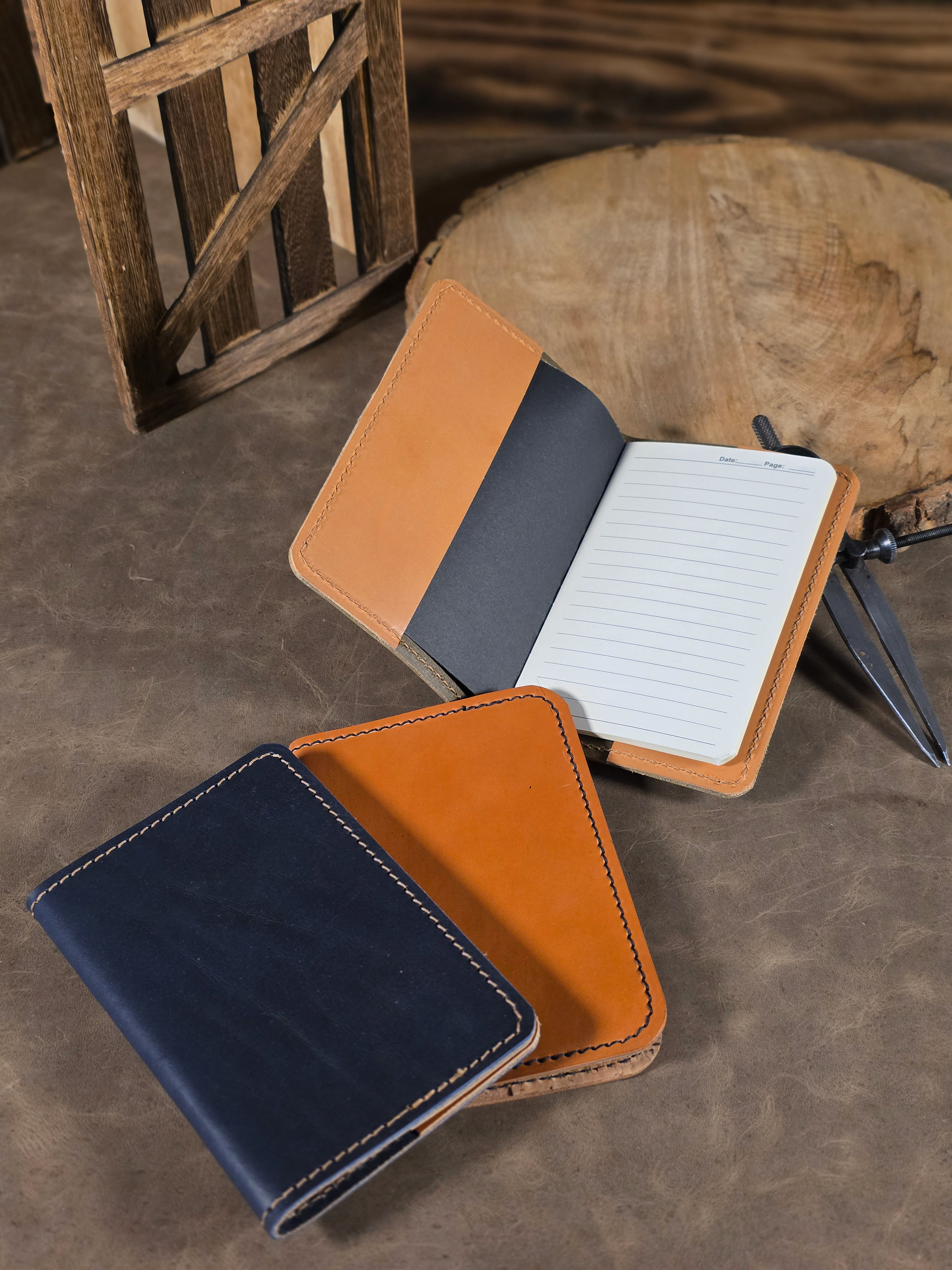 Three leather notebook journal covers sitting on a vintage leather mat. One journal is navy leather and the other is made of russet leather. One of the journals is sitting open where the interior russet leather is visible as it leans against a wooden slab.