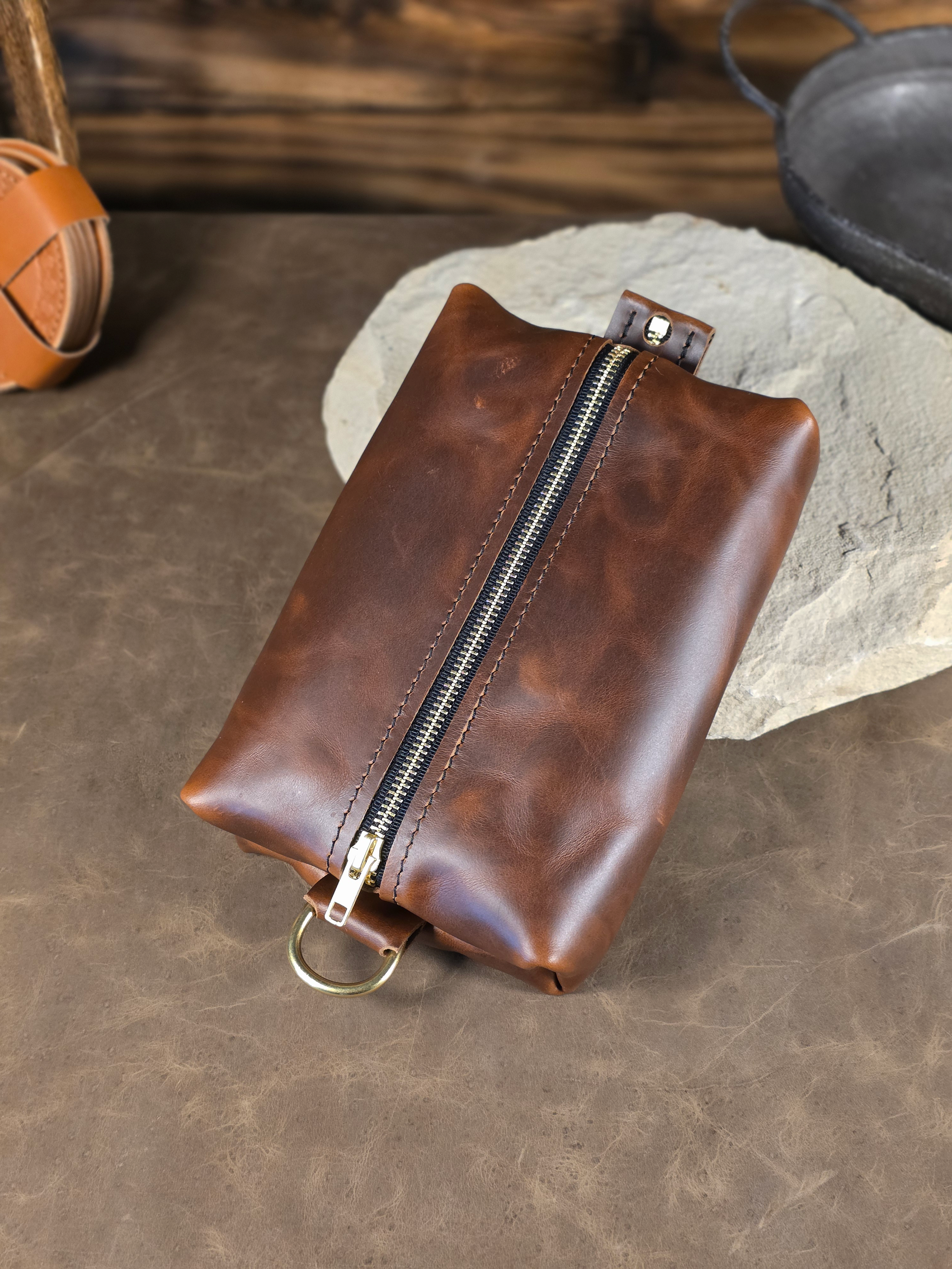 Brown leather dopp kit pouch with a solid brass zipper resting on a pale stone surface