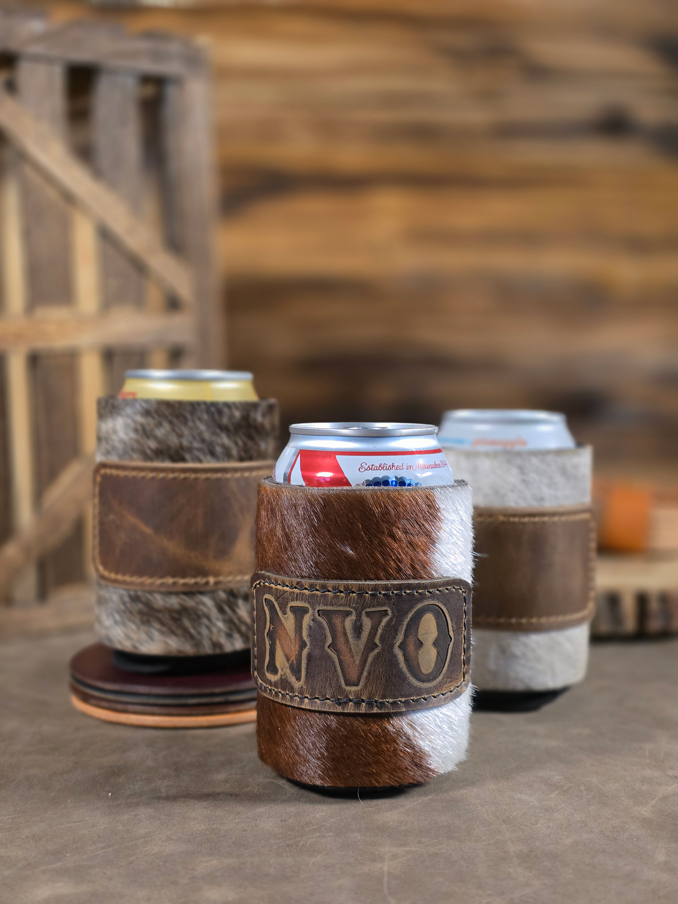 Three hair-on leather cowhides koozies sitting in a rustic background, each koozie has a rustic brown leather patch. The middle koozie has the initials NVO hot branded onto the leather.