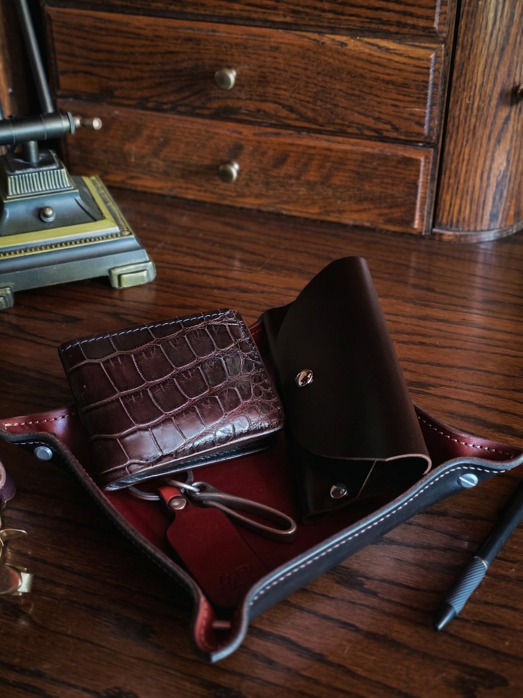 Brown leather valet tray holding an alligator wallet and dark brown leather sunglass case on a wooden surface with a vintage lamp in the background