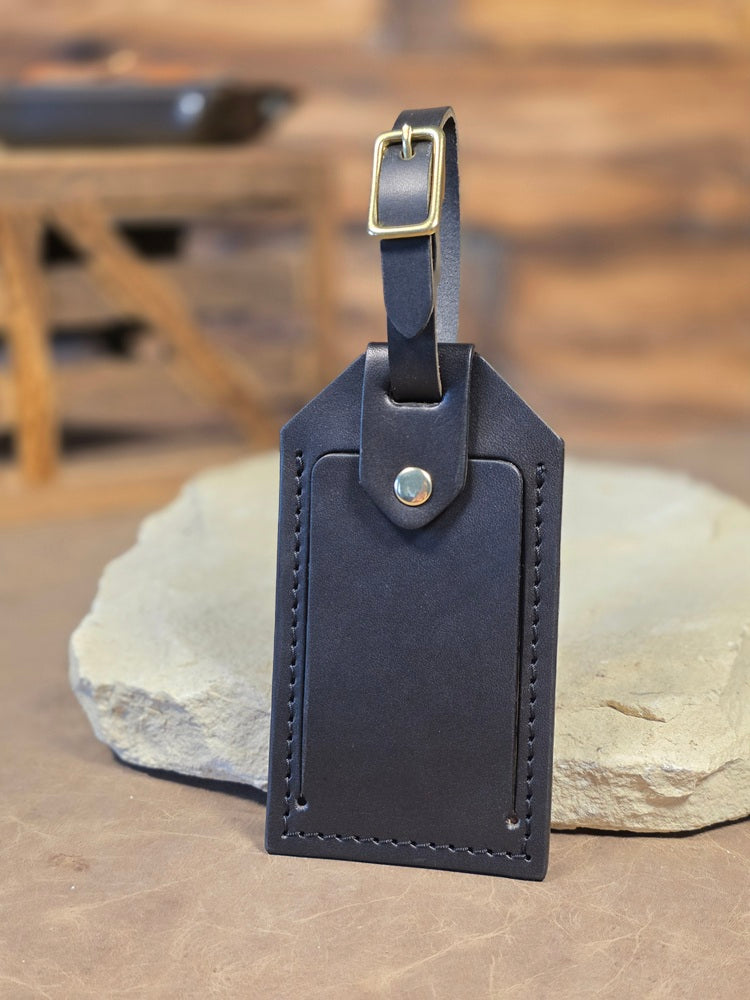 Close up view of a black leather windowed luggage tag finished with solid brass hardware and black stitching. The luggage tag leans against a white rock.