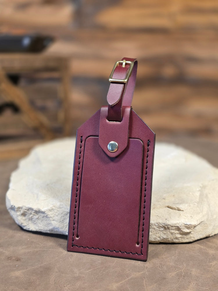 Close up view of a oxblood leather windowed luggage tag finished with solid brass hardware and black stitching. The luggage tag leans against a white rock.
