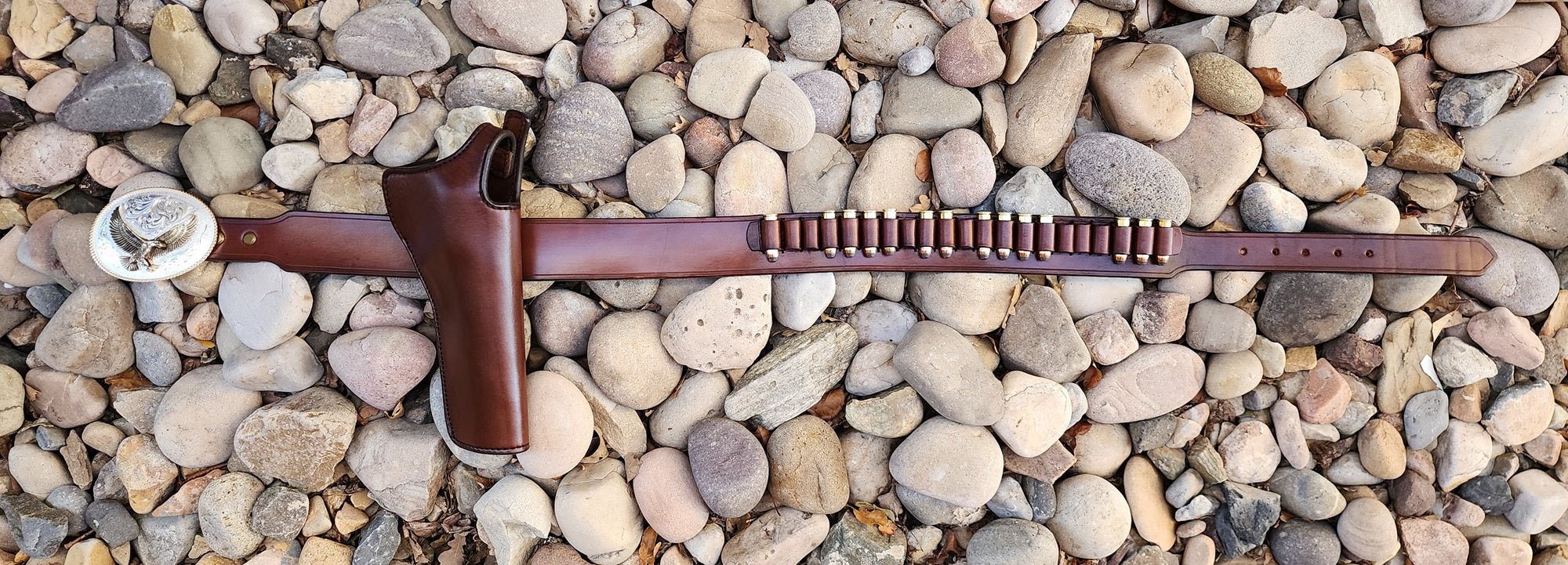 Full view of the Lone Wolf Mcquade gun rig fully layed out on a bed of pale river rocks. The buckle is silver and gold on the left side of the rig, next to it is the holster made of brown leather just like the belt. Close to the billet end of the belt are 21 bullet loops, 14 of which are filled.