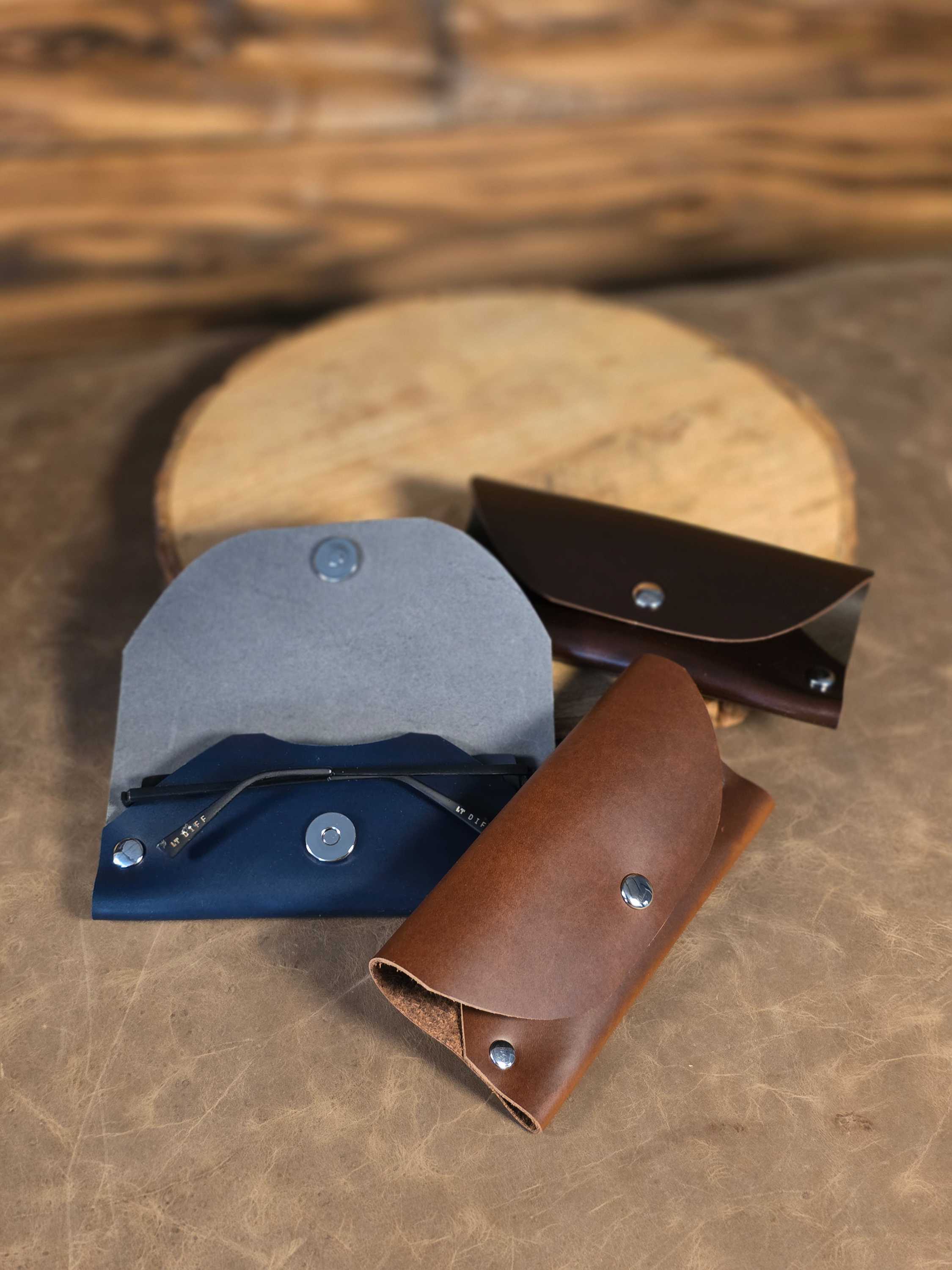 Three soft leather sunglass cases sit on a wooden table, they are blue, chocolate and cognac colored.