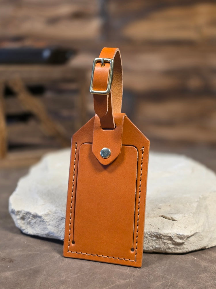 Close up view of a russet leather windowed luggage tag finished with solid brass hardware and tan stitching. The luggage tag leans against a white rock.