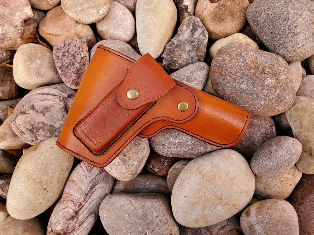 Russet leather vintage 1911 holster with flap coverage, on the flap is a magazine pouch. The holster is finished with solid brass hardware and black stitching, the piece lays on a bed of pale river rocks.