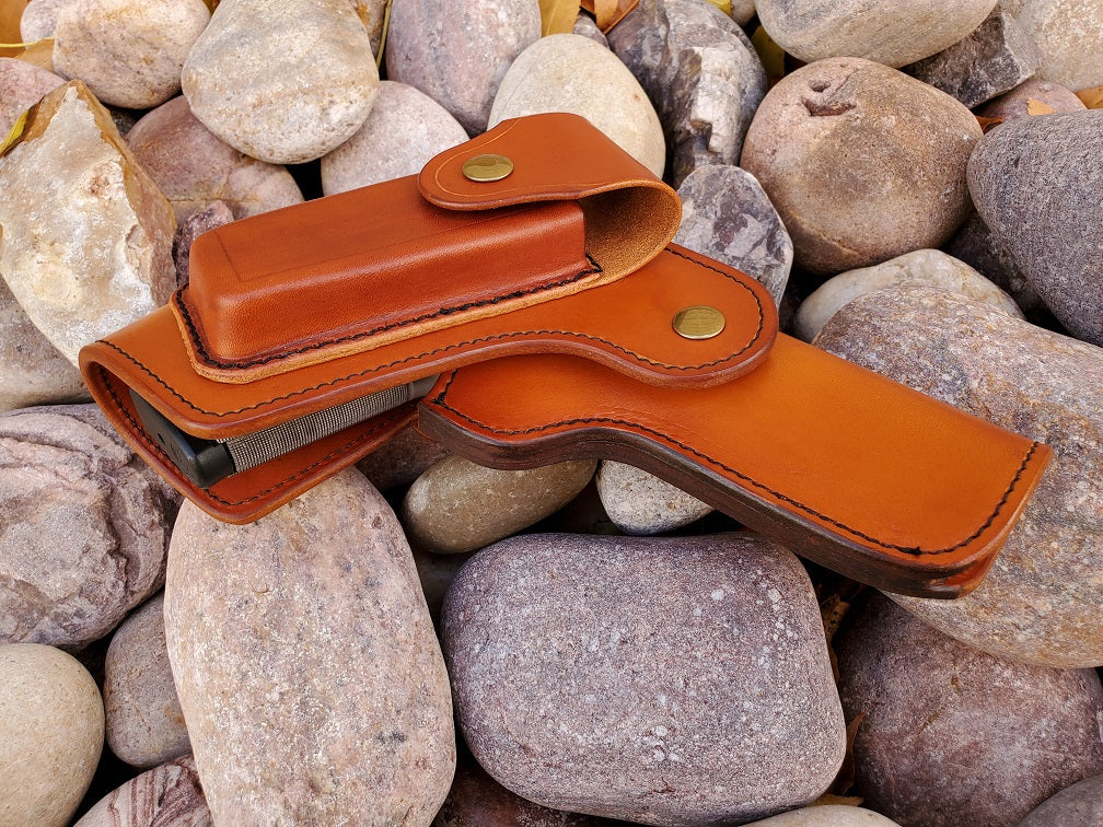 Side view of the russet leather vintage 1911 holster with flap coverage, on the flap is a magazine pouch. The holster is finished with solid brass hardware and black stitching, the piece lays on a bed of pale river rocks.