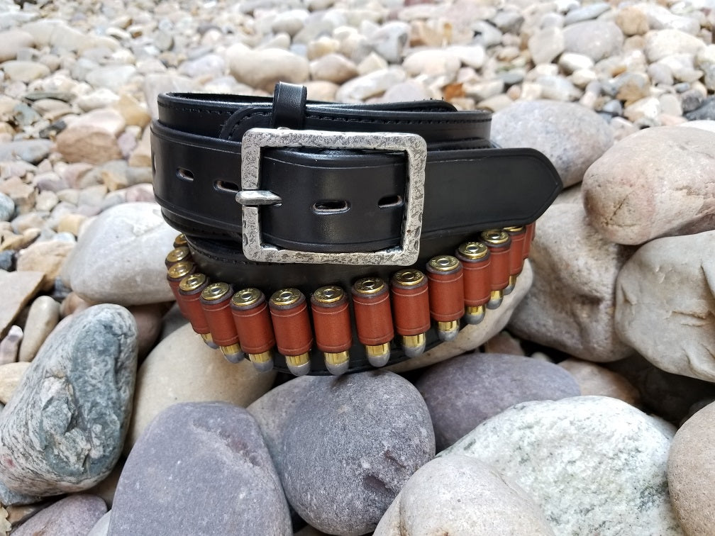 Black cowboy belt with hammered buckle coiled around itself sitting on smooth river rocks.