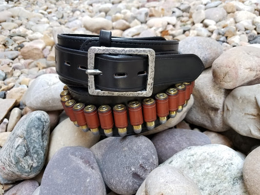 Black cowboy belt with hammered buckle coiled around itself sitting on smooth river rocks. 