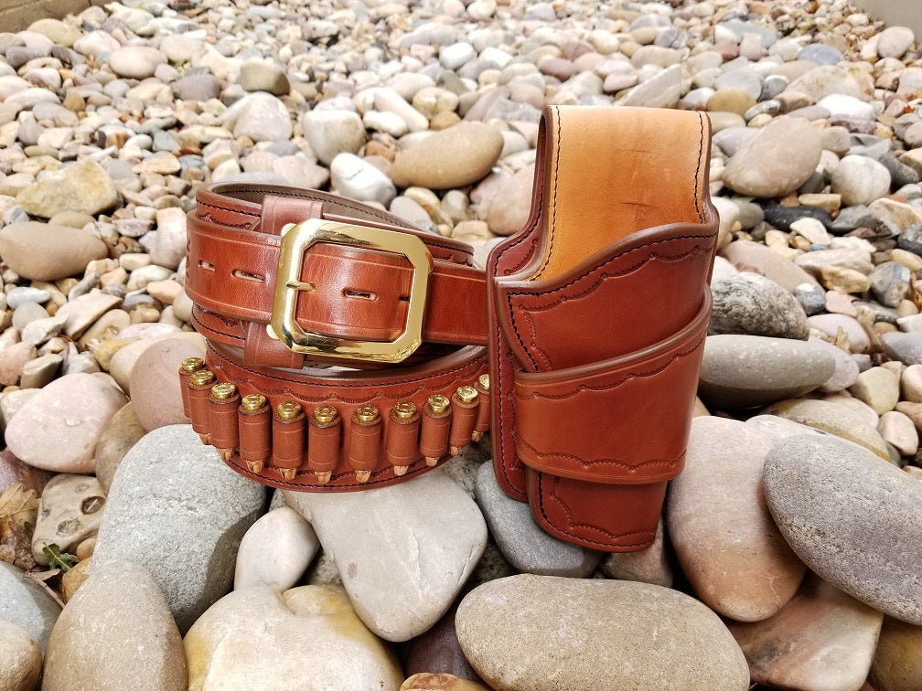 Widow rock gunbelt and holster. The rig is made of brown leather tooled with a scallop pattern border. Additionaly, the hardware on the rig is made of brass. The rig sits on top of a bed of pale river rocks.