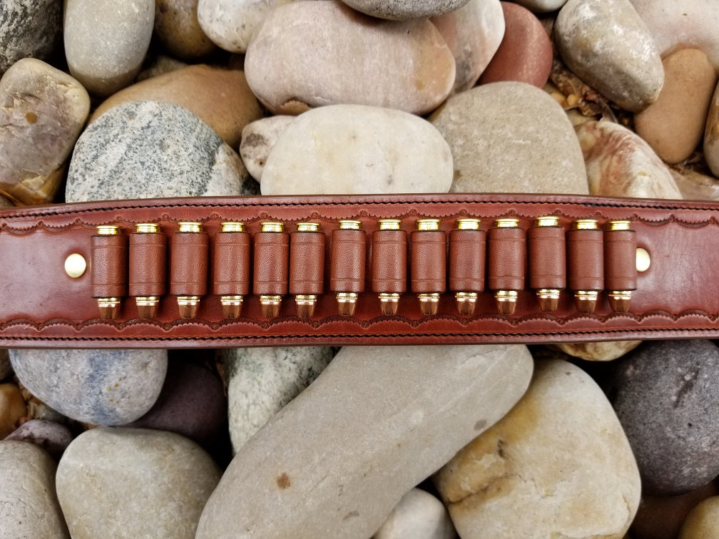 Section of widow rock belt featuring bullet loops sitting on a bed of pale river rocks. The piece is made of brown leather and solid brass hardware with scallop pattern tooling along the borders.