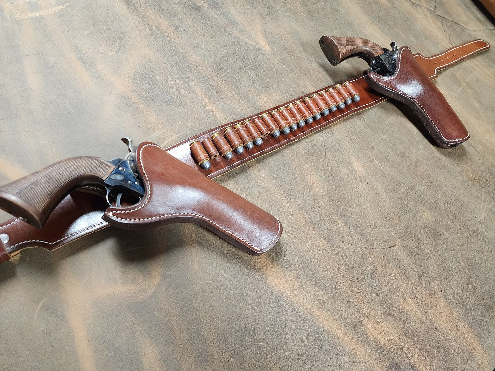 View of a brown leather gun rig stretching to the left, pictured is a strong draw and cross draw holster with 18 bullet loops, the piece sits on a ghost leather bed.