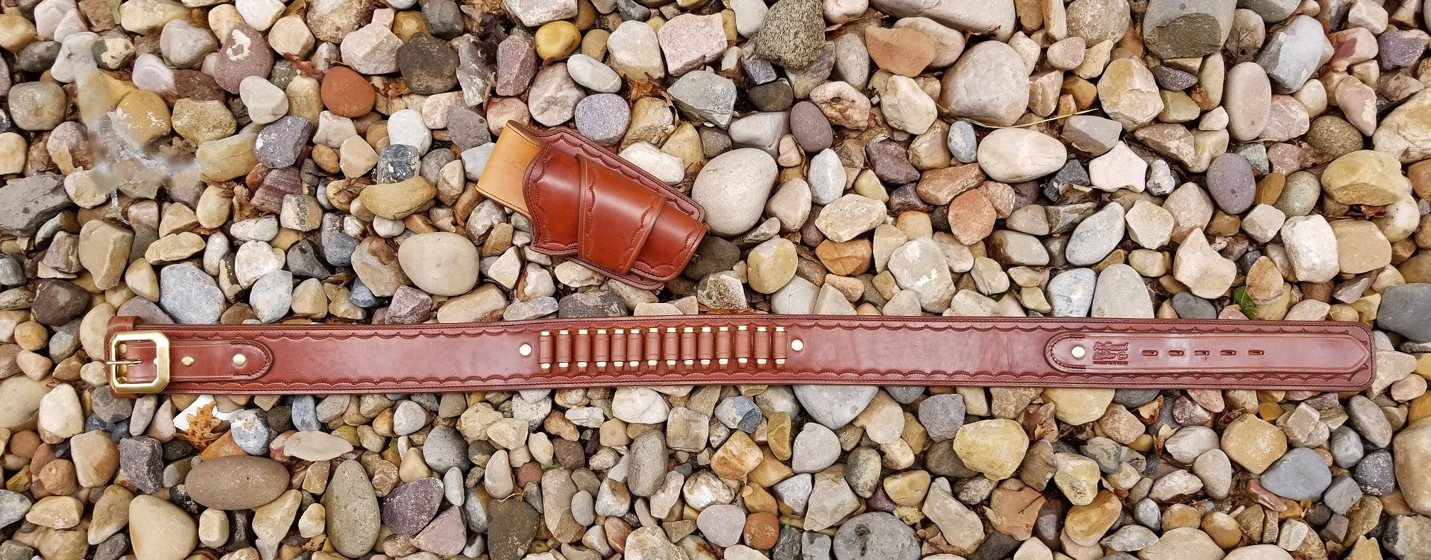 Widow rock gun rig layed out across a bed of rocks. The gun rig is made of brown leather and solid brass hardware, the piece is tooled with a scallop pattern along the border.