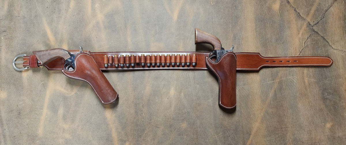 Josh Faraday inspired gun rig with right hand strong arm draw and right handed cross draw holsters attached to a brown leather belt with eighteen bullet loops. The piece is finished with white stitching and solid brass nickel hardware. The piece sits on a bed of ghostwood leather.