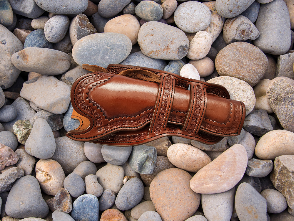 Brown leather holster which is intricately tooled with a double Mexican loop collar. The holster is sitting is a bed of pale river rocks.