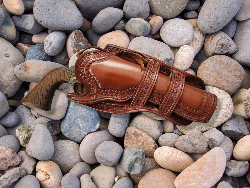 Brown leather holster which is intricately tooled with a double Mexican loop collar and holstering a revolver. The holster is sitting is a bed of pale river rocks.