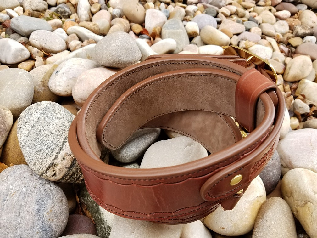 Widow rock belt sitting on a bed of pale stones. The piece is made of brown leather and solid brass hardware with scallop pattern tooling along the border.