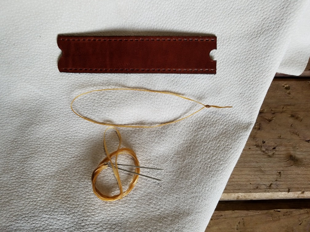 unassembled leather wrap placed on white leather next to sinew pieces with two needles.