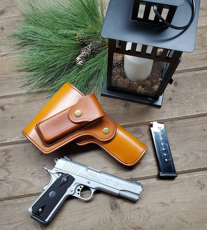 Russet leather vintage 1911 holster with full flap coverage and a magazine pouch attached to the flap, next to the holster is a 1911 and a 1911 magazine, all the items lay on wooden slats.