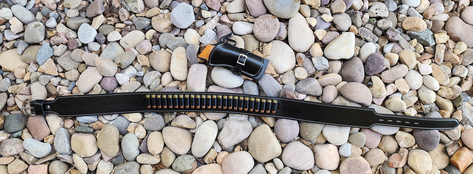 Angel eyes gun rig fully layed out on a bed of pale river rocks, the holster is not attached. The holster and belt are made of black leather and finished with white stitching and solid brass nickle plate hardware, the belt is additionaly finished with twenty four bullet loops.