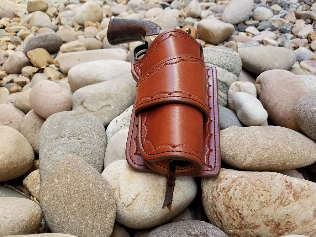 Brown leather holster sitting on a bed of rocks, the holster is tooled in a scallop pattern. Holstered is a colt revolver.