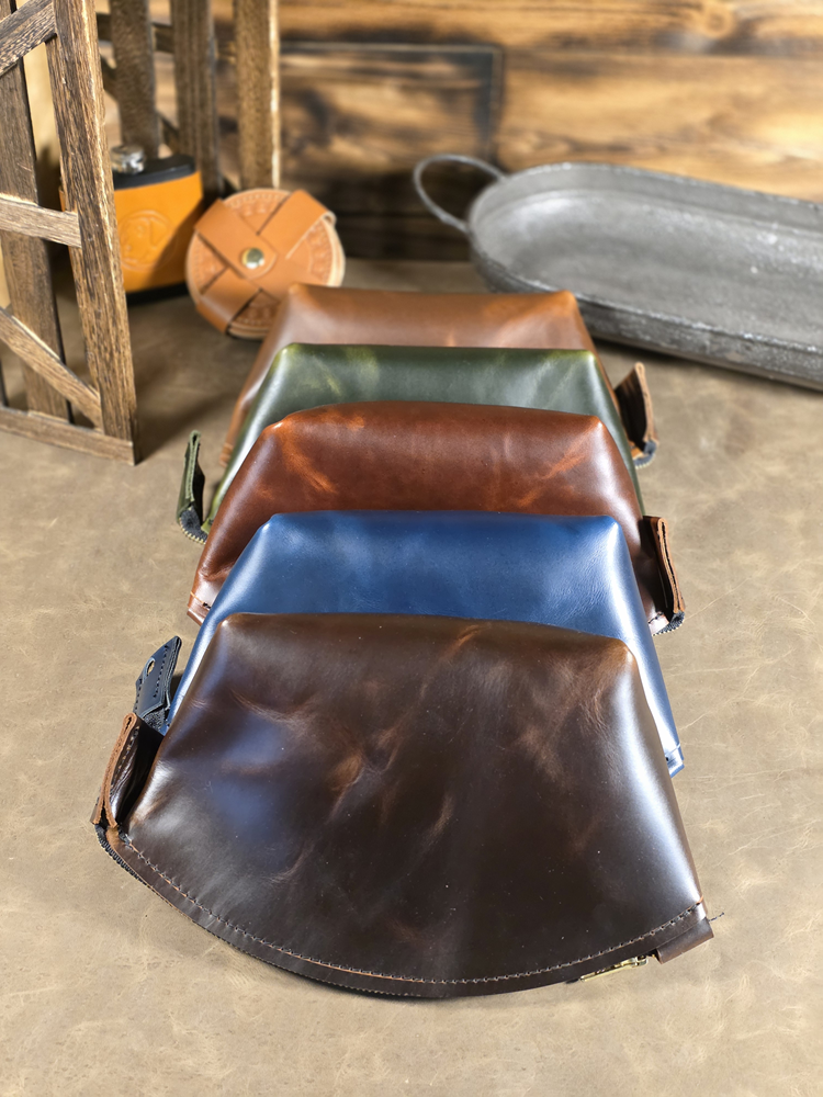Five leather cosmetics pouches in chocolate, navy, antique copper, forest green and cognac colors on a wooden surface