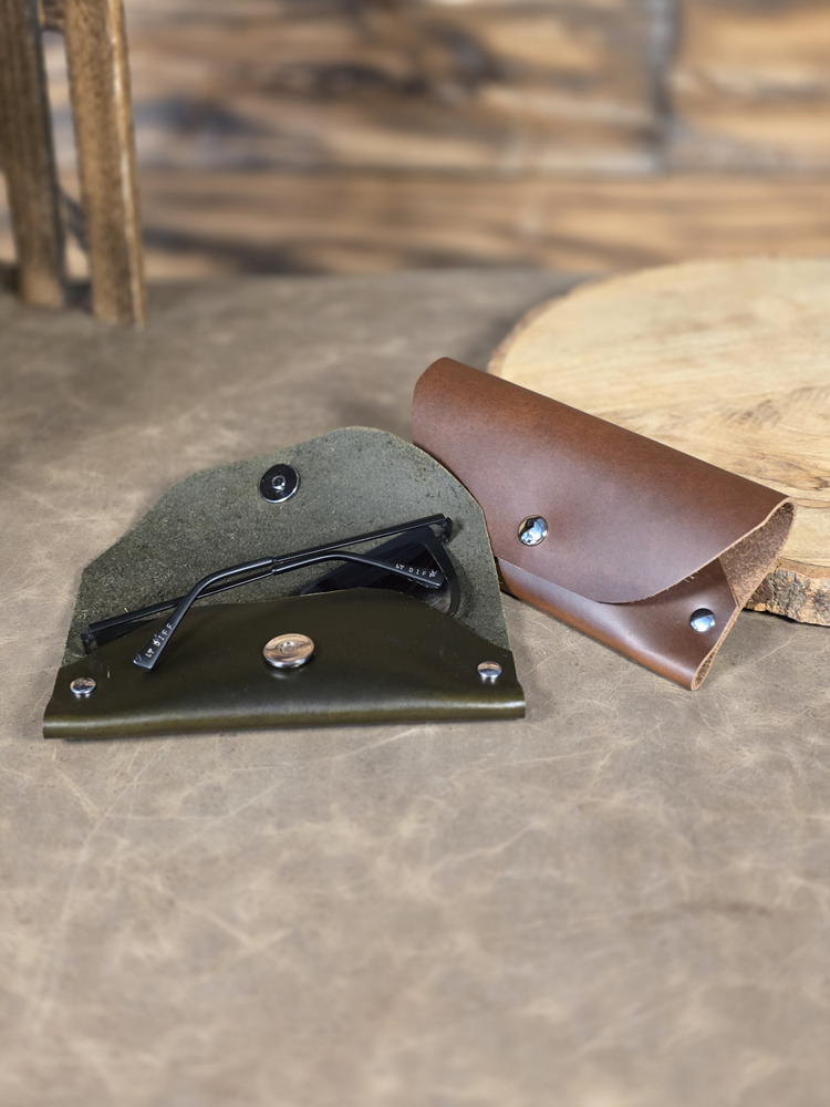 Two leather sunglass pouches, one green and one brown, on a textured surface. The green sunglass pouch is open, showing the sunglasses inside.