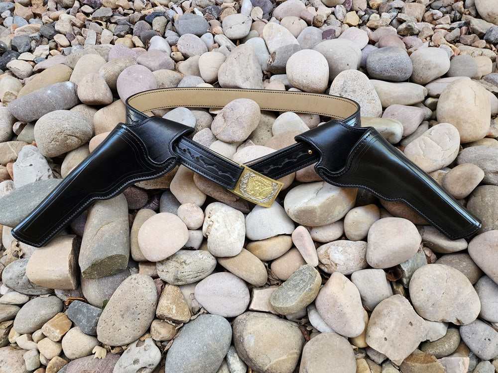 Sitting on a bed of pale stones is a black leather replica of Wild Bill Hickok's gun rig, featuring two matching holsters and a classic silver and gold cavalry officers buckle.