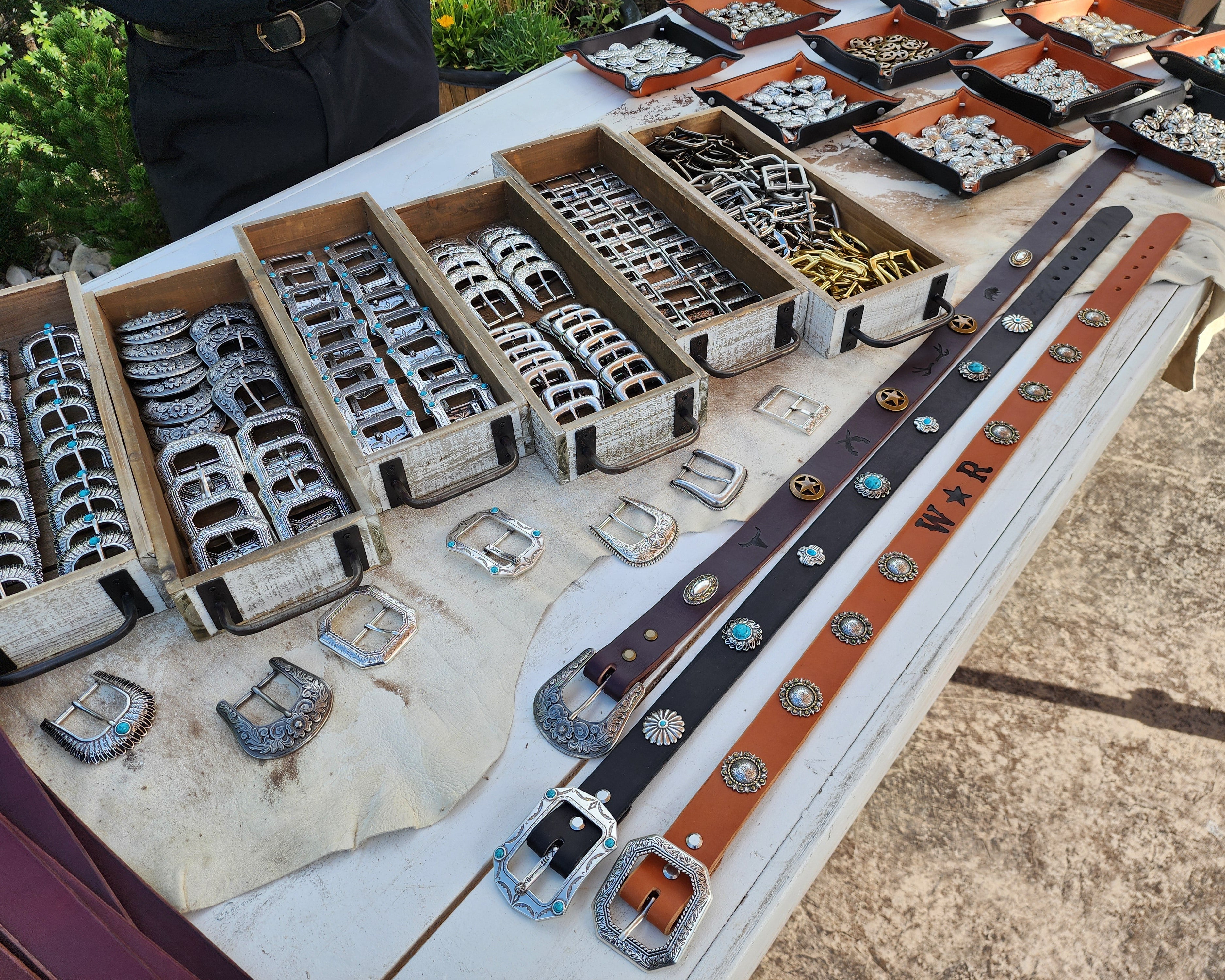 Several wooden and leather trays contain a large variety of beautiful western style buckles and conchos. Nearby, a series of beautifully decorated and hot branded sample belts lay across a wooden farm table.