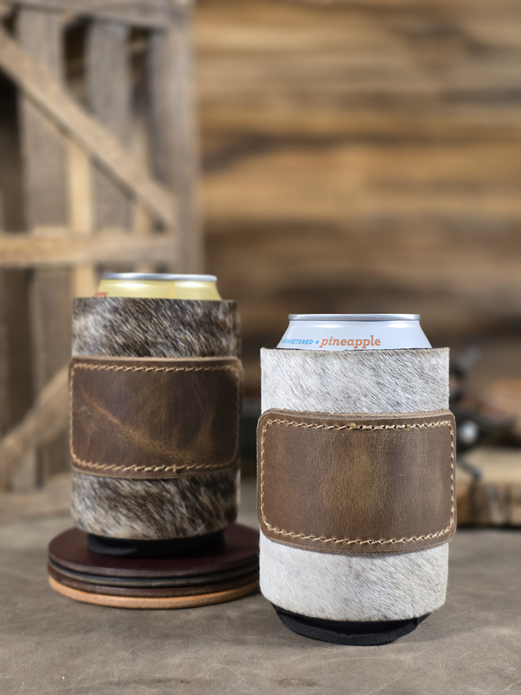 Two hair-on cowhide koozies sitting on a rustic background, each koozie has a rustic brown patch.