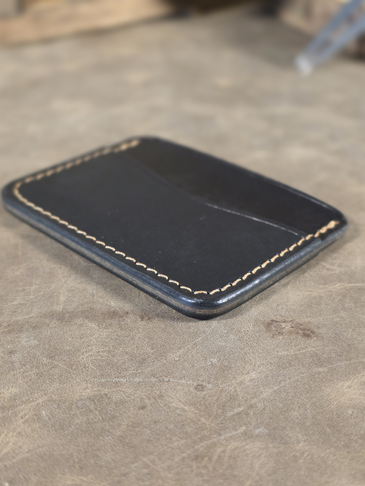 All black leather minimalist wallet, focus on slicked hand burnished edges with tan stitching on a gray surface