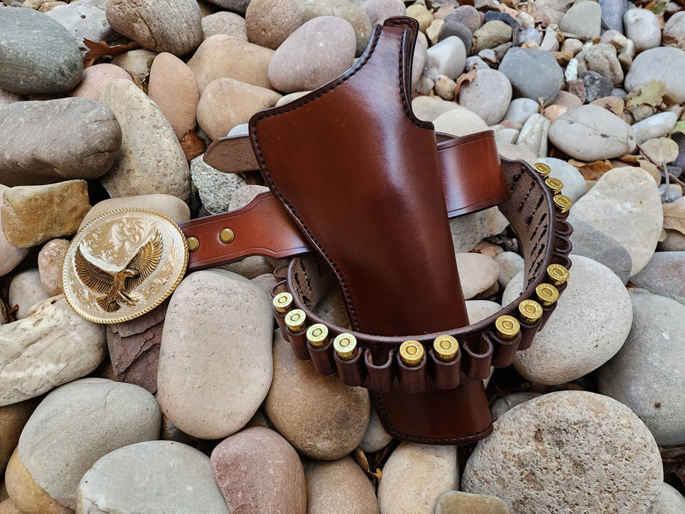 Brown leather gun belt and holster with some bullet loops filled out, the image focuses on the holster with the silver and gold plated buckle just to the left of the holster. The rig lays on a bed of river rocks.
