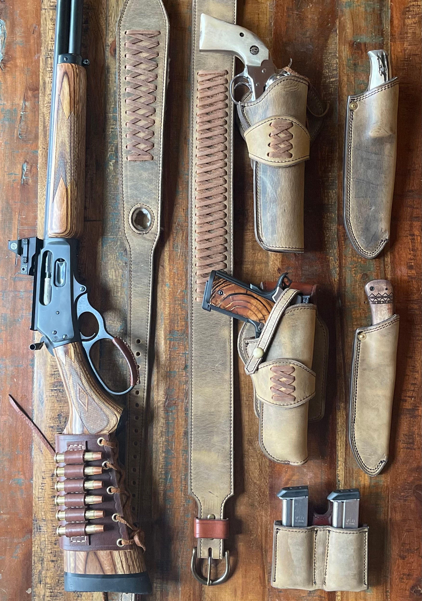A variety of Hellhound's signature Ghostwood gunleather pieces displayed on a wooden table. Near a rifle sits a Ghostwood thumbhole sling, a Ghostwood scout belt, a matching double mag pouch, two sheaths and the Classic Ghostwood Holsters