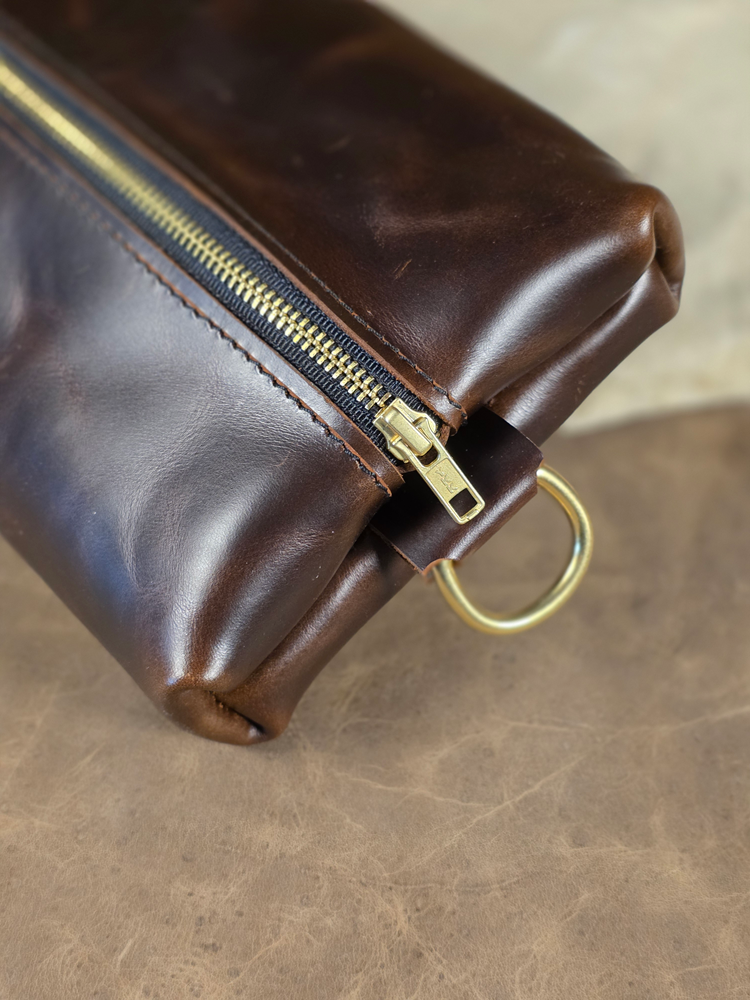 Brown leather dopp pouch with gold zipper and D ring on a matching background