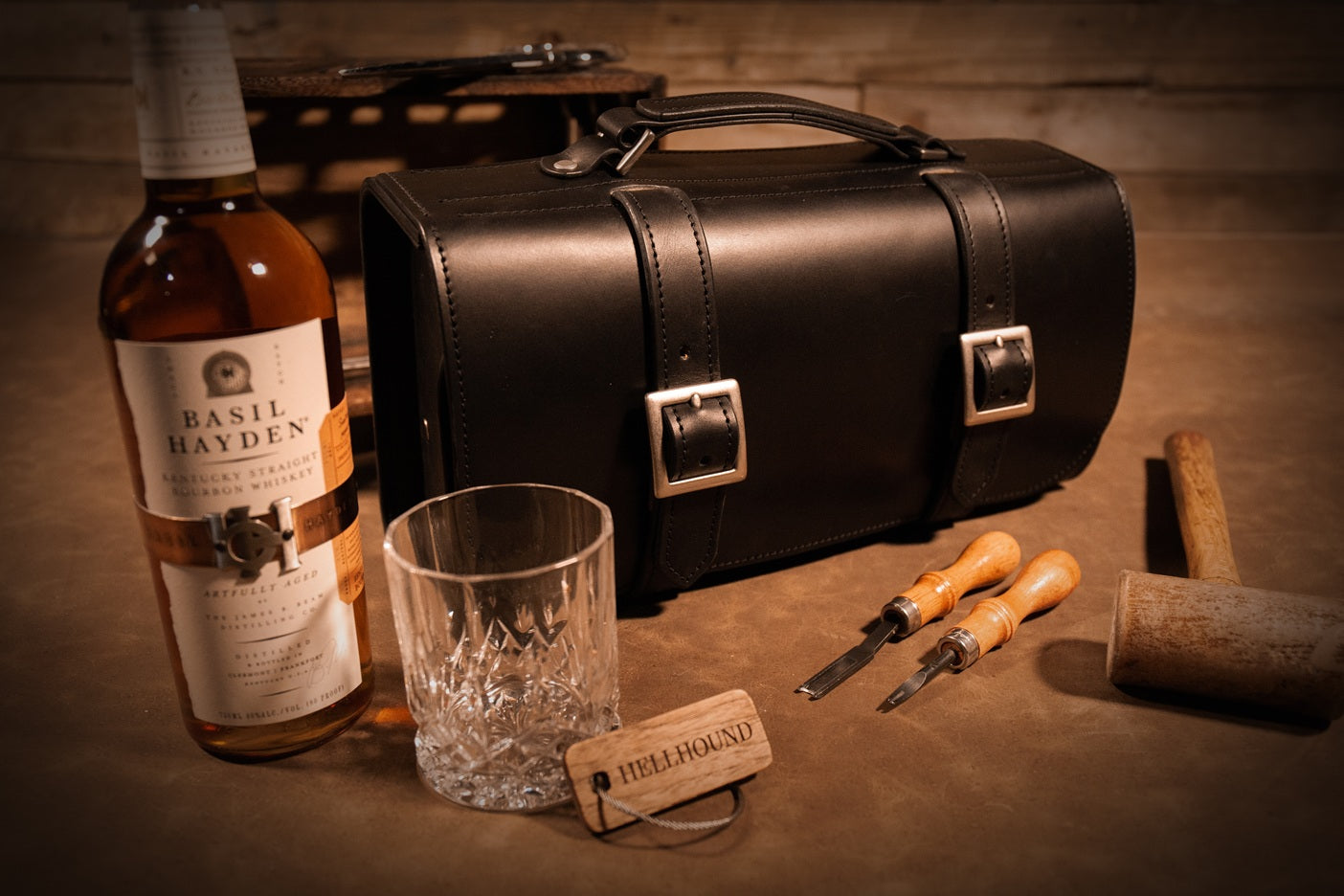 Bottle of Basil Hayden whiskey, black  English Bridle leather bourbon bag, a rocks glass, and leathercrafting tools on a wooden surface.
