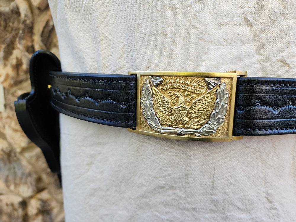 Close up of the buckle on replica of Wild Bill Hickok's belt, the belt is black leather and tooled in a scallop pattern, the buckle is a classic US army cavalry buckle in gold and silver colors.