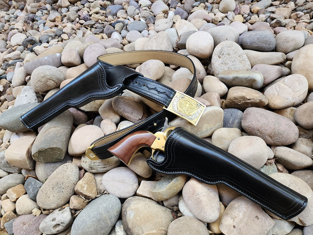 Sitting on a bed of pale rocks, a black leather replica of Wild Bill Hickok's gun rig is displayed. The rig features two holsters, one holster is attached to the belt, which features a classic silver and gold cavalry buckle. The other holster sits just beside the belt and is holstering a 1873 Colt Peacemaker.